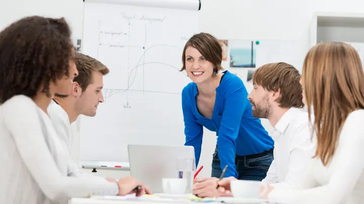 Eine lächelnde Person steht vor einem Flipchart auf dem eine Grafik zu sehen ist und spricht mit ihrem Team aus vier Personen.