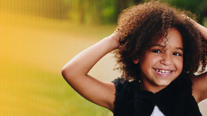 Ein fröhliches Kind mit dunklen Locken und einer Zahnlücke greift sich in die Haare und lächelt in die Kamera.