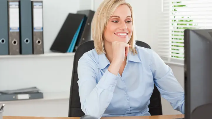 Eine blonde junge Frau sitzt lächelnd vor ihrem Bildschirm im Hintergrund sieht man Ordner in einem Regal.